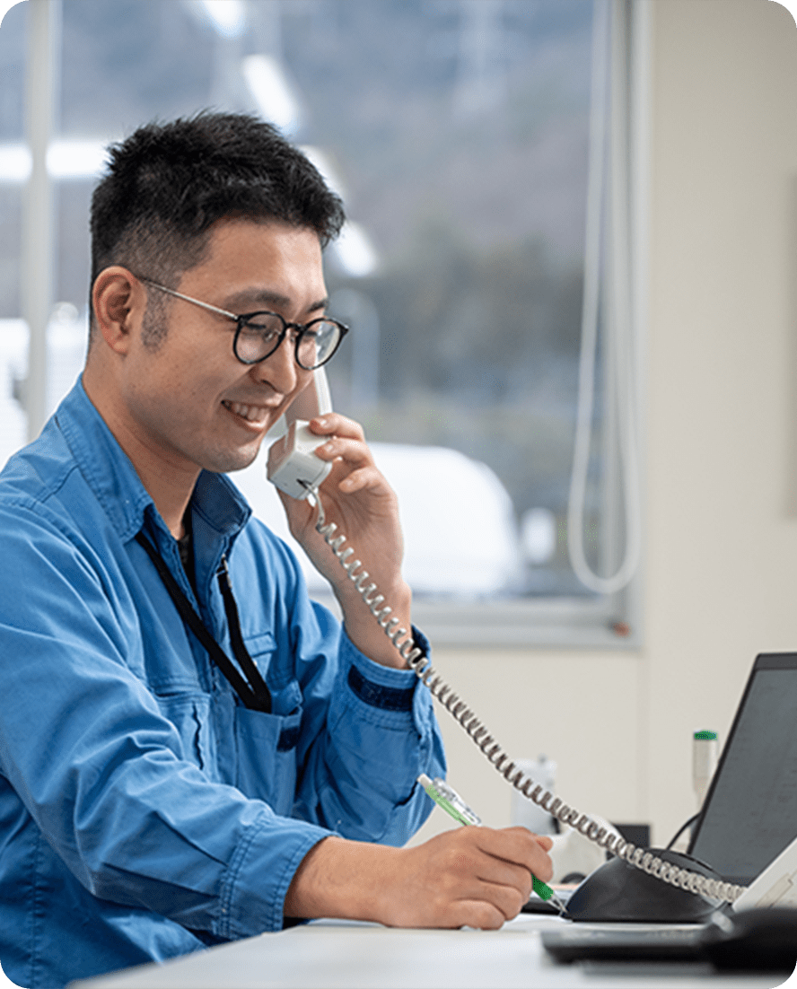 青い作業服を着た男性が、オフィスで電話をしながらメモを取っている様子