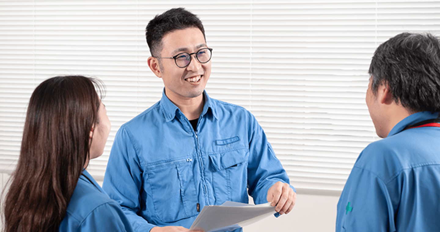 青い作業服を着た3人の社員がオフィスで会話をしている様子