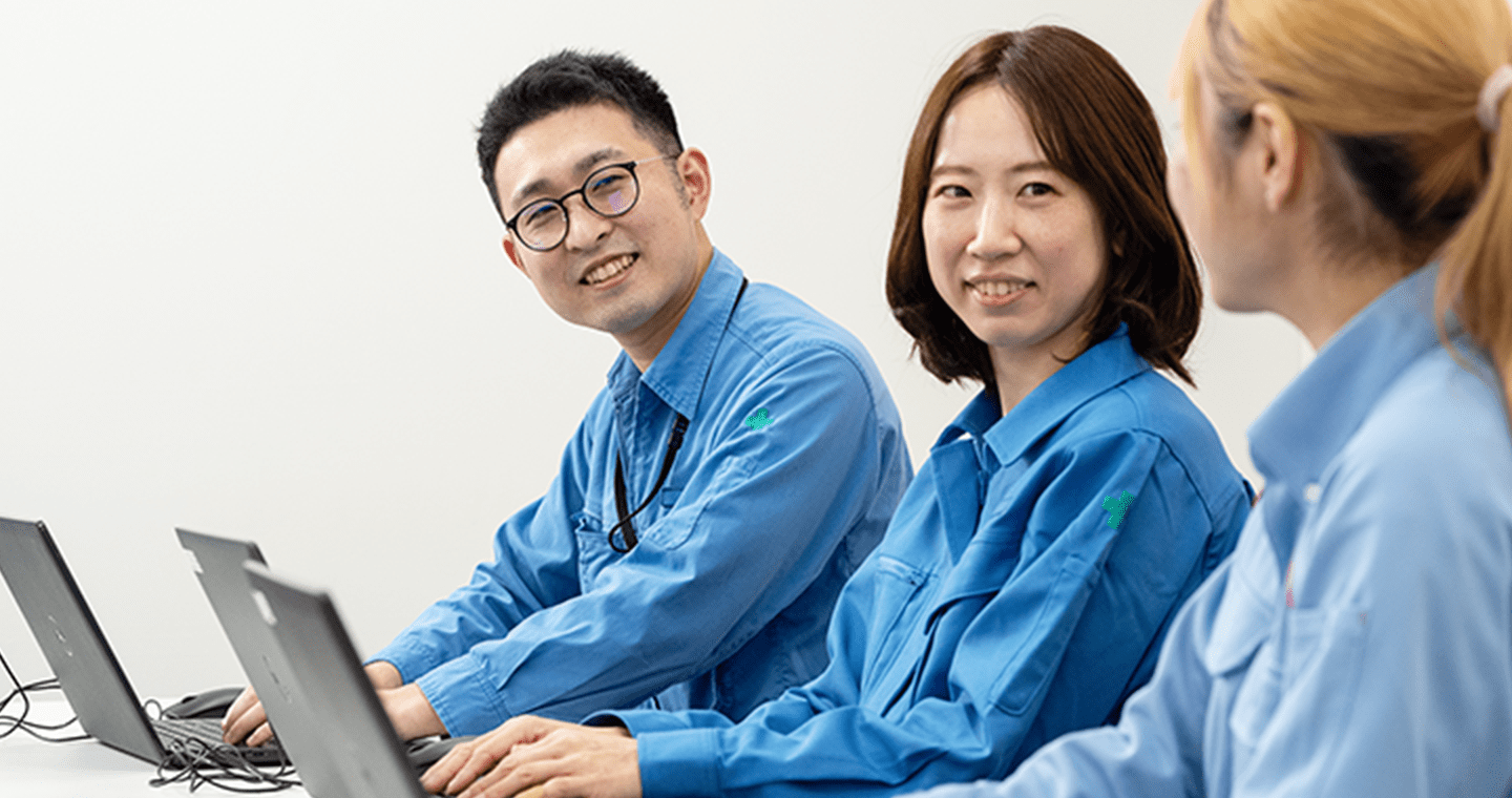 青い作業服を着た3人の社員が、ノートパソコンを使いながら会話をしている様子