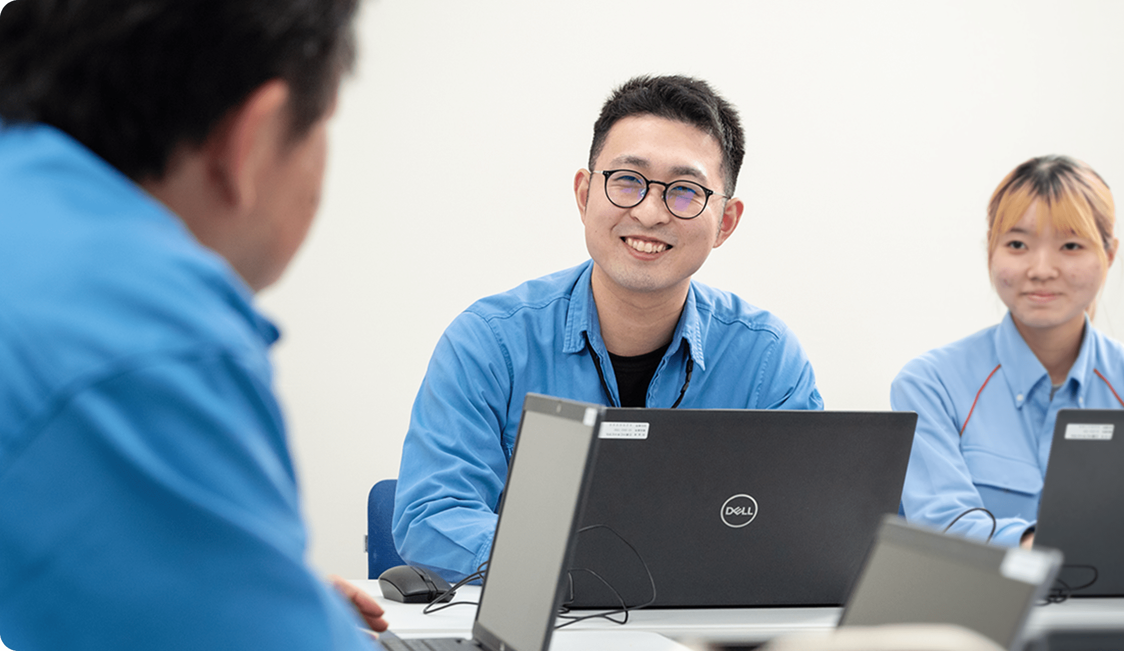 青い作業服を着た社員3人が会議室でノートパソコンを使いながら打ち合わせをしている様子