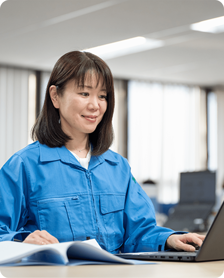 青い作業服を着た女性が、オフィスでノートパソコンを使って作業している様子