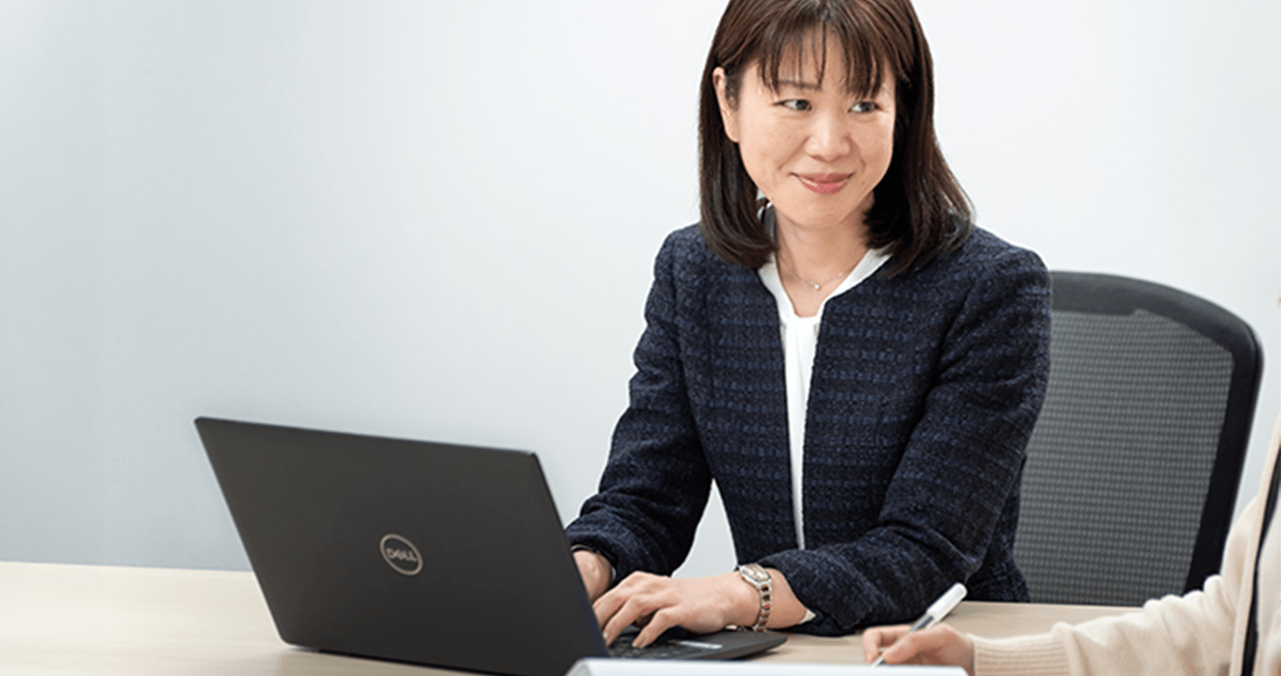 黒いジャケットを着た女性がノートパソコンを操作しながら、向かいの相手と会話している様子