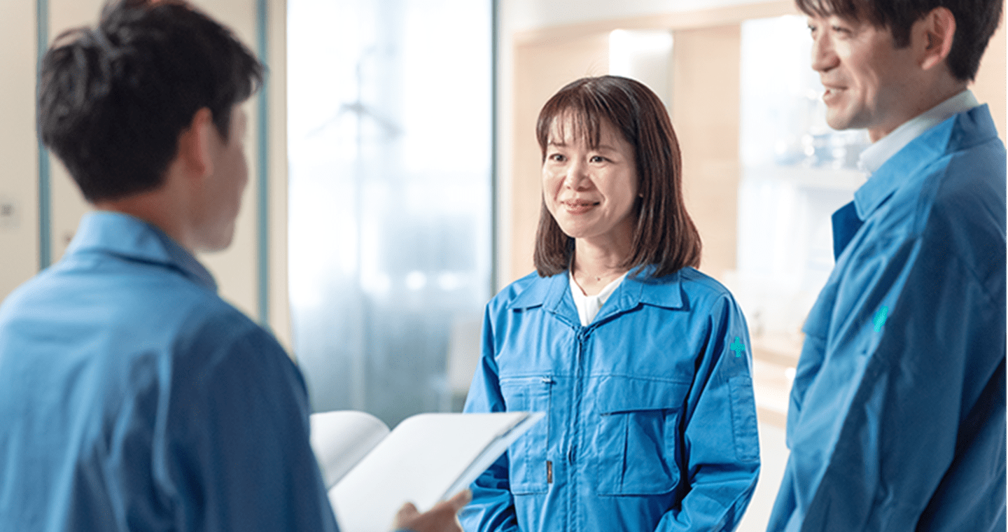 青い作業服を着た3人の社員が、職場で打ち合わせをしている様子