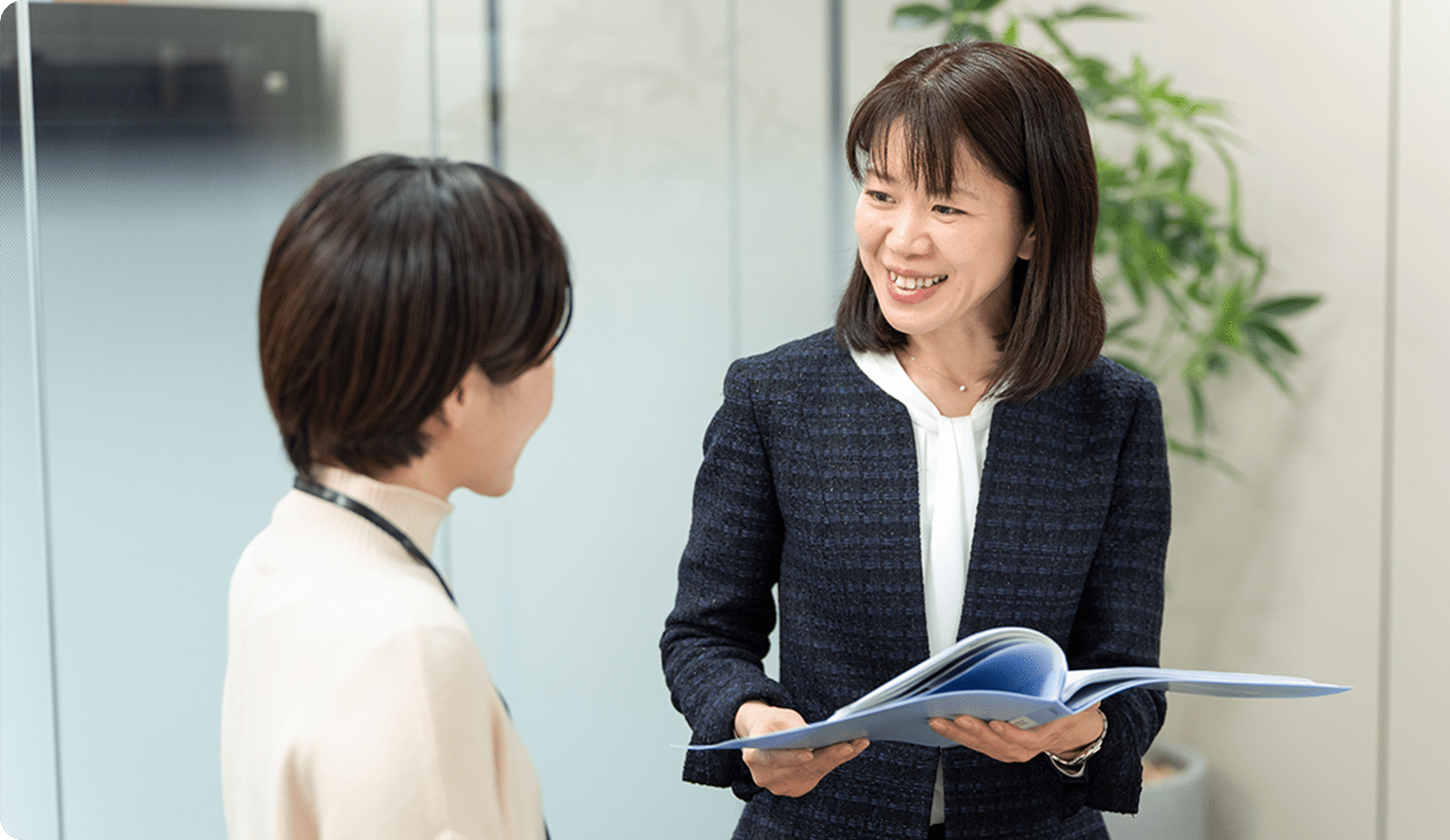 黒いジャケットを着た女性が、書類を持ちながら笑顔で話をしている様子