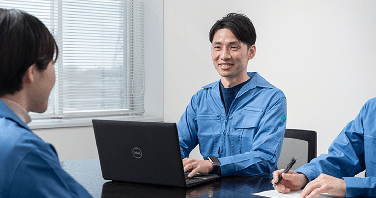 青い作業服を着た男性が、ノートパソコンを前にして笑顔で会話している様子