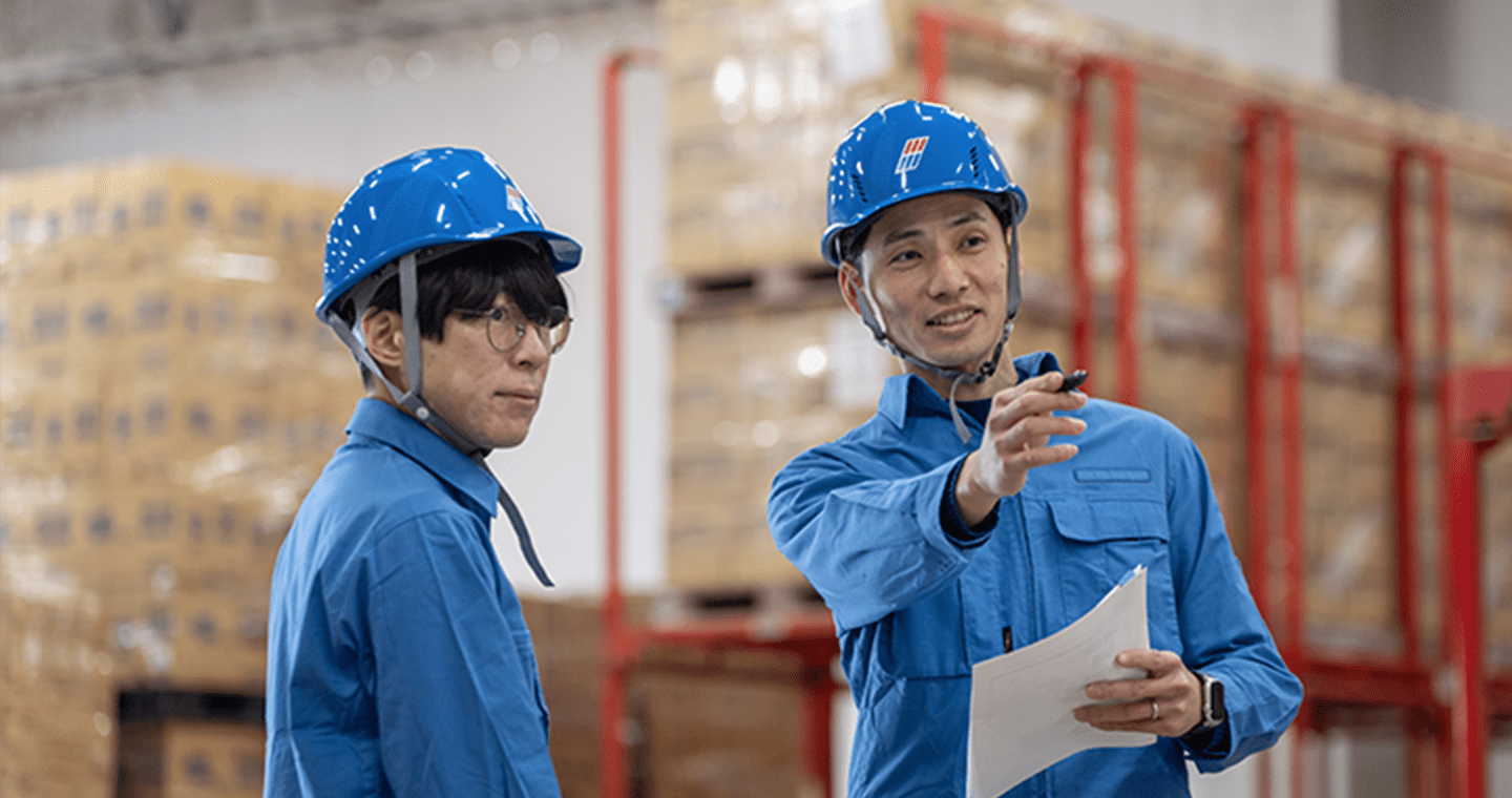 青い作業服とヘルメットを着用した2人の作業員が、倉庫内で打ち合わせをしている様子