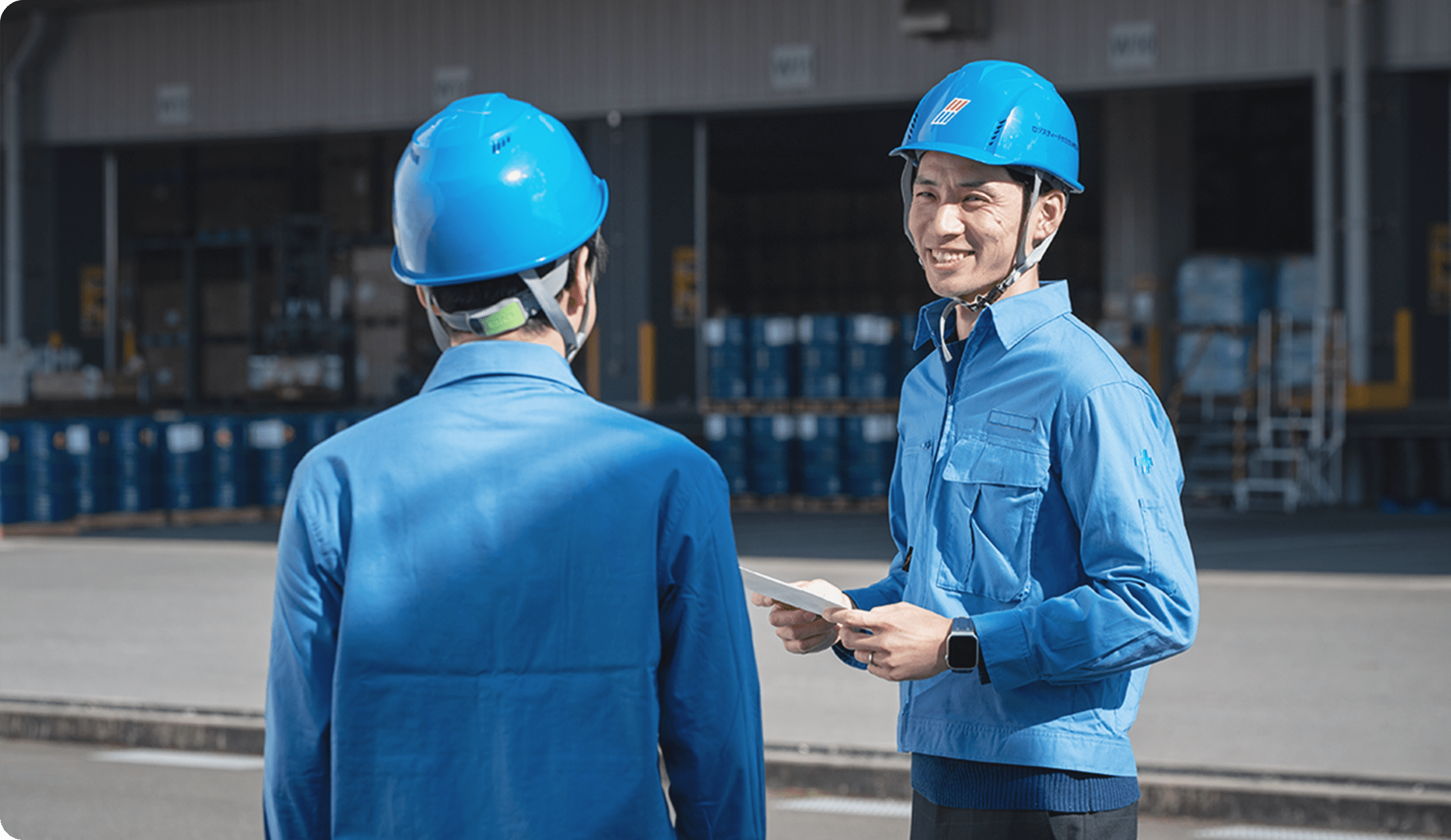 青い作業服とヘルメットを着た2人の作業員が、屋外の倉庫前で会話をしている様子