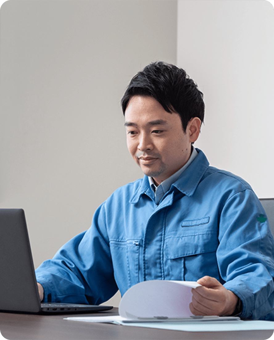 青い作業服を着た男性が、ノートパソコンと書類を使いながら作業している様子