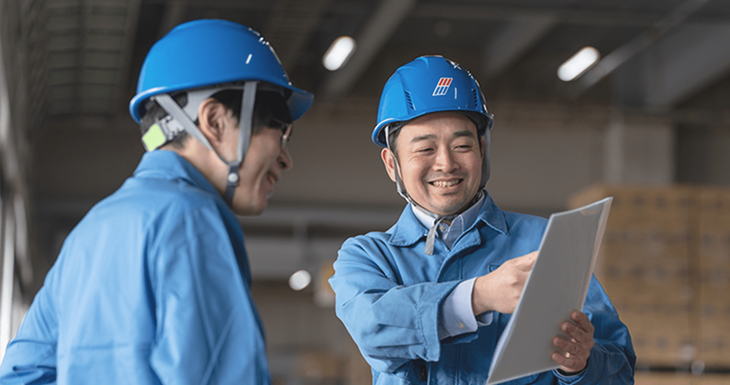 青いヘルメットと作業服を着た2人の作業員が、倉庫内で書類を確認しながら会話している様子