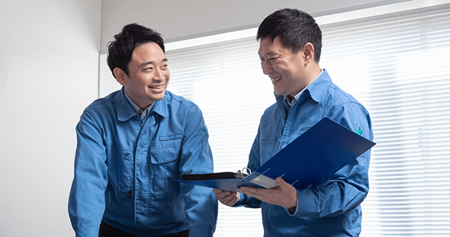 青い作業服を着た2人の男性が、オフィスで書類を見ながら笑顔で話し合っている様子