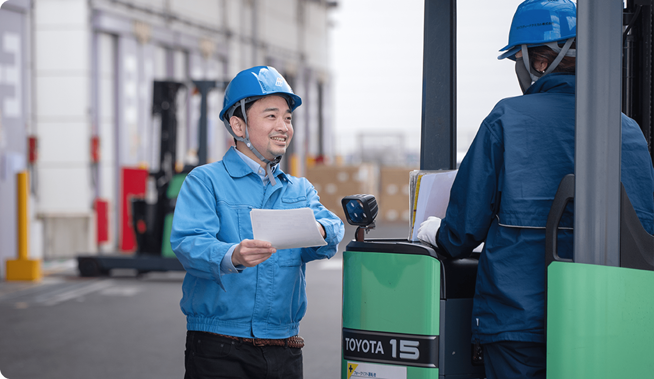青いヘルメットと作業服を着た男性が、フォークリフトの運転手に書類を手渡している様子