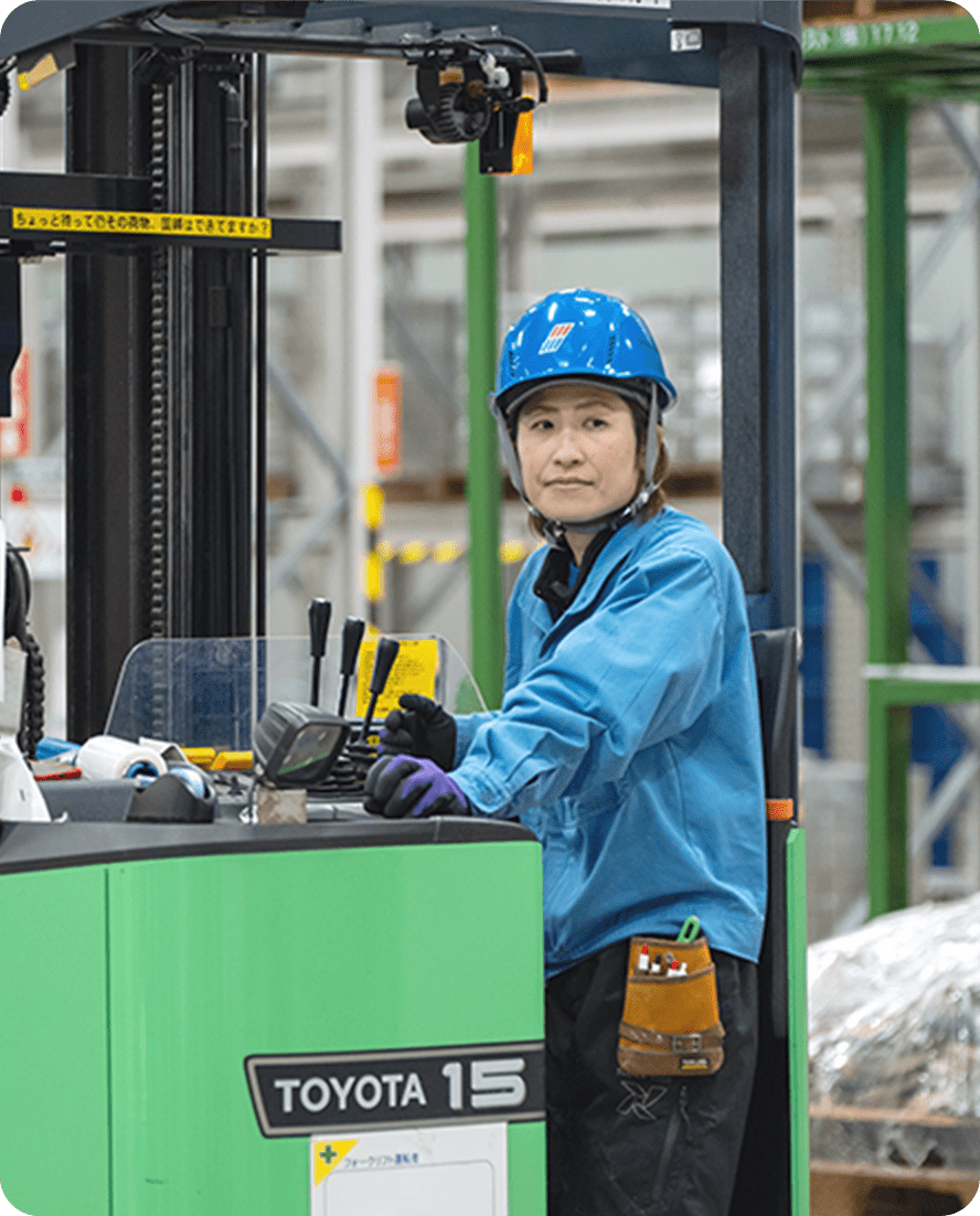 青い作業服とヘルメットを着た女性が、倉庫内でフォークリフトを操作している様子
