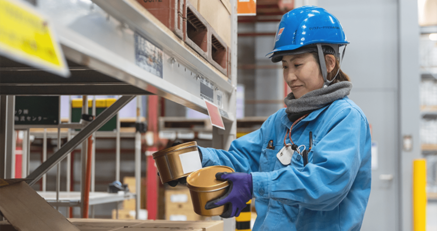 青い作業服とヘルメットを着た女性が、倉庫の棚から金属製の容器を取り出している様子