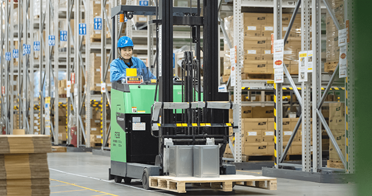 青い作業服とヘルメットを着た女性が、倉庫内でフォークリフトを運転し荷物を運搬している様子