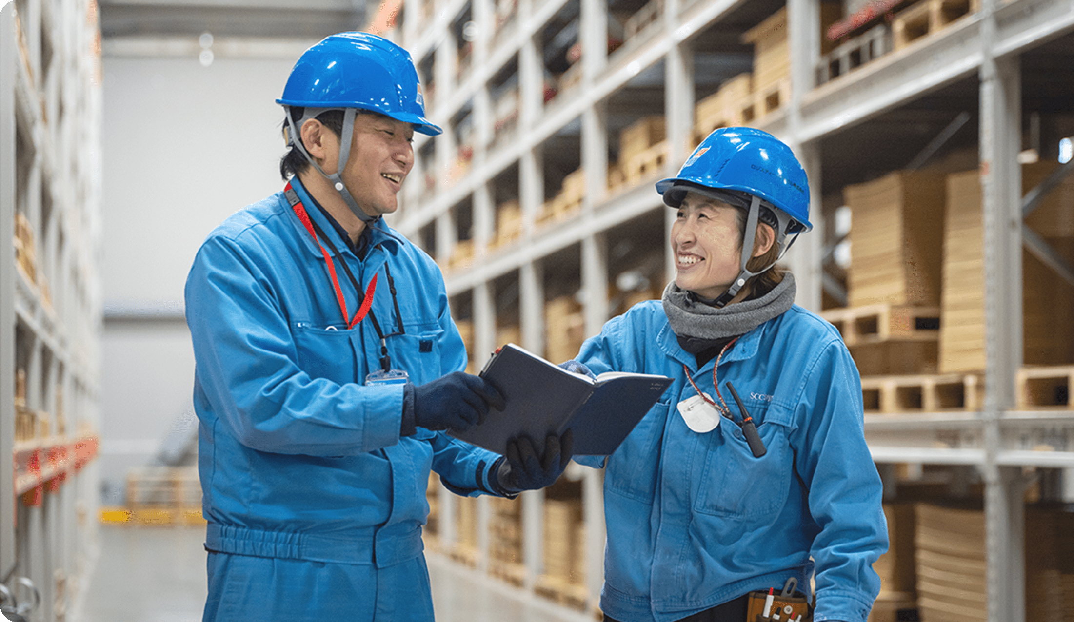 青い作業服とヘルメットを着た男女が、倉庫内で笑顔で話し合いながら打ち合わせをしている様子