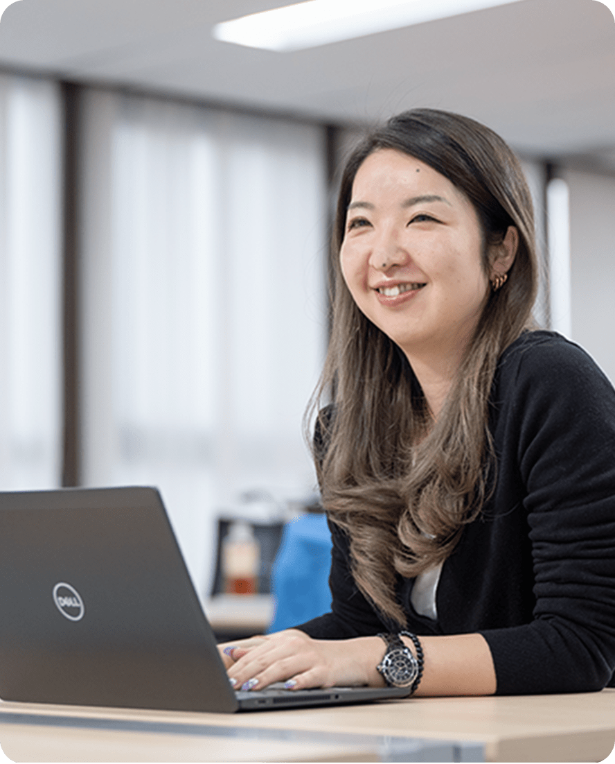 黒いカーディガンを着た女性が、オフィスでノートパソコンを使いながら微笑んでいる様子