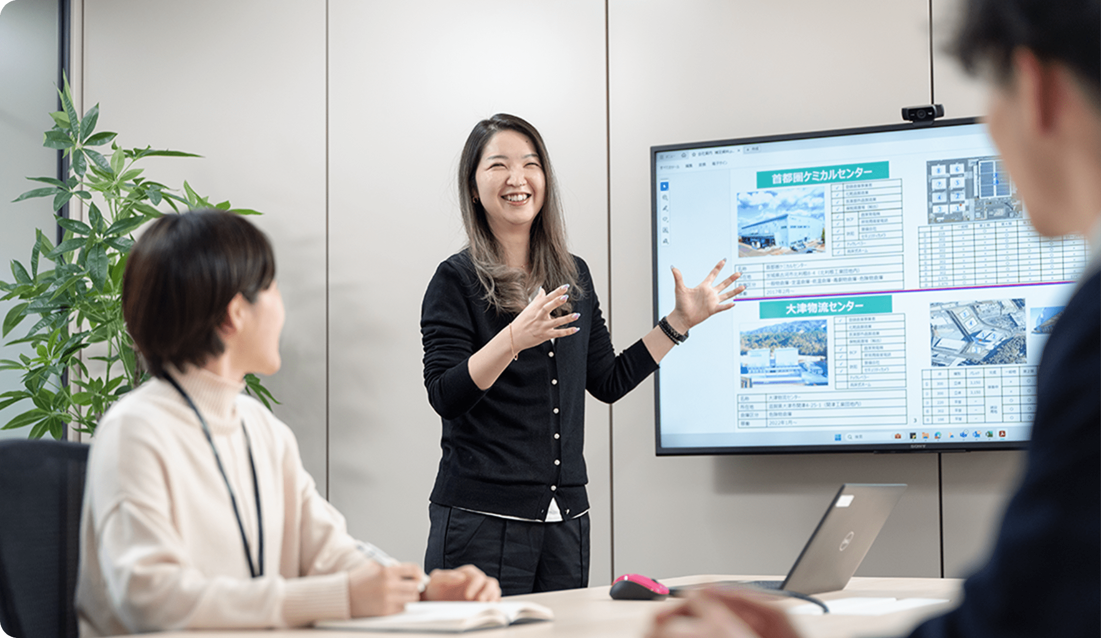 会議室で黒いカーディガンを着た女性が、プレゼンテーションを行っている様子