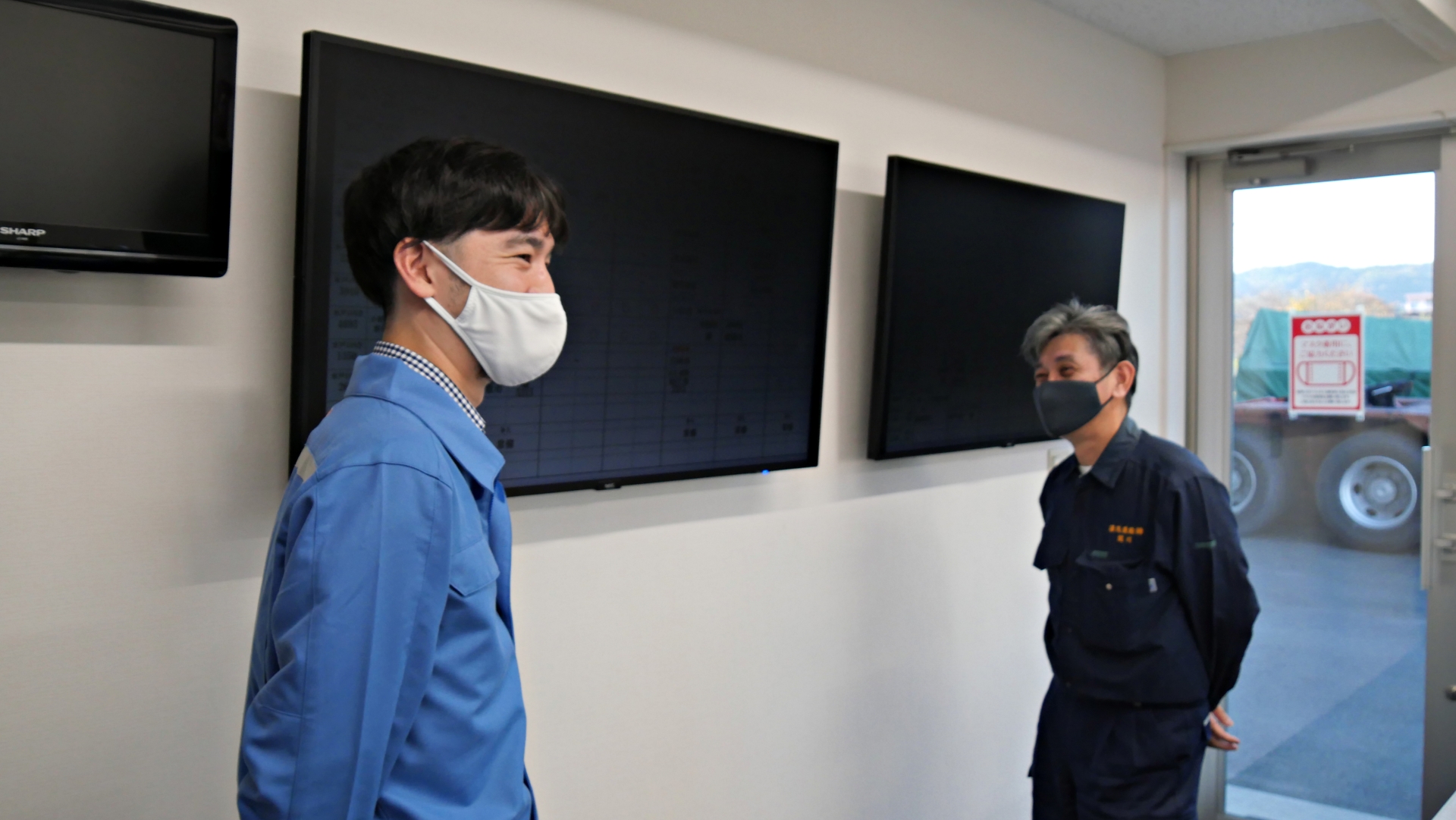 湯浅運輸の事務所内には、配車表を表示するためのモニターが2台設置されている。モニターの前で談笑する関川氏(右)と日立物流 輸送事業強化PJ SSCV強化グループ主任 服部哲郎氏(左)
